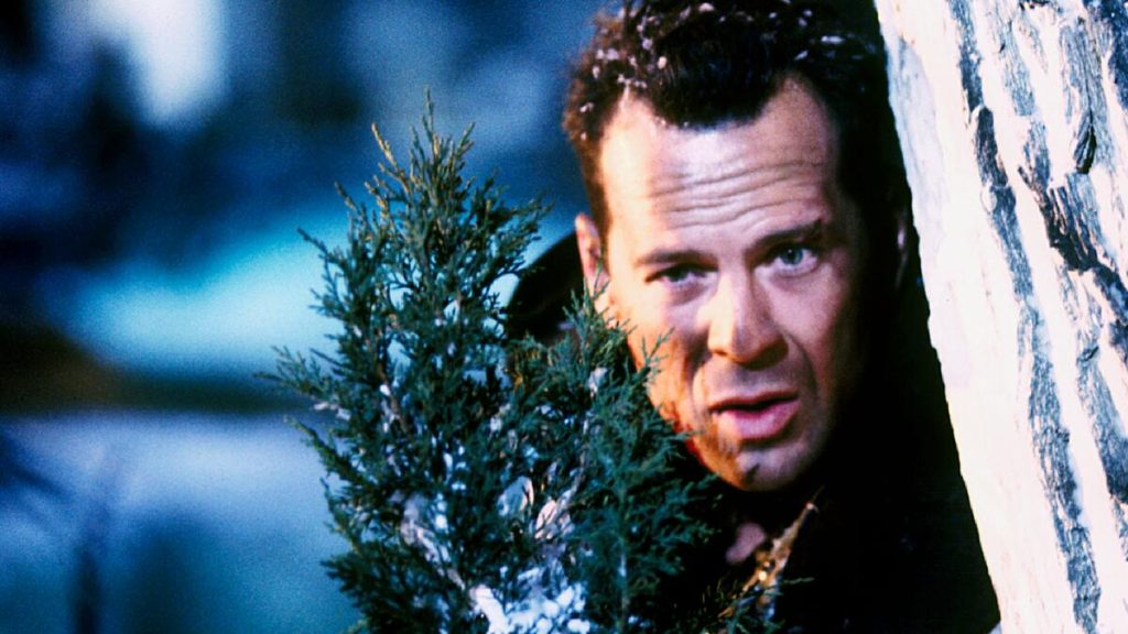 A still image from the movie Die Hard 2, featuring Bruce Willis’ character holding a small Christmas tree.